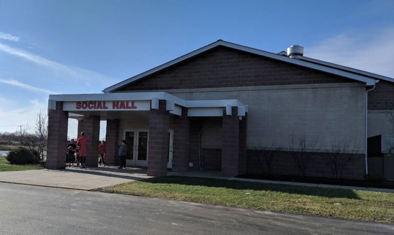 A group of people stands near the entrance of a one-story building labeled "SOCIAL HALL" in red letters, with a clear sky and sunlight casting shadows on the ground.